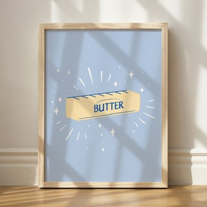 May include: A framed print with a light blue background featuring a stick of butter with the word "BUTTER" printed on it. The butter is surrounded by white stars and lines.