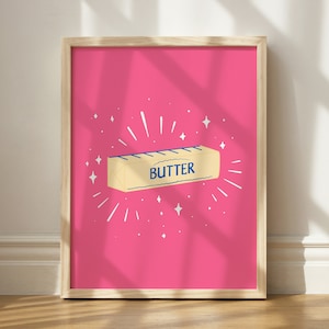 May include: A framed print with a pink background and a cartoon illustration of a stick of butter with the word "BUTTER" printed on it. The butter is surrounded by white stars and white lines radiating outward.