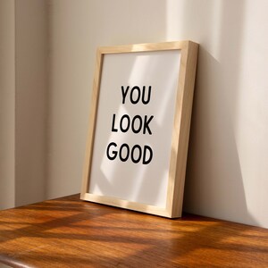 May include: A framed print with the text "YOU LOOK GOOD" in black lettering on a white background. The frame is made of light wood and is sitting on a wooden surface.