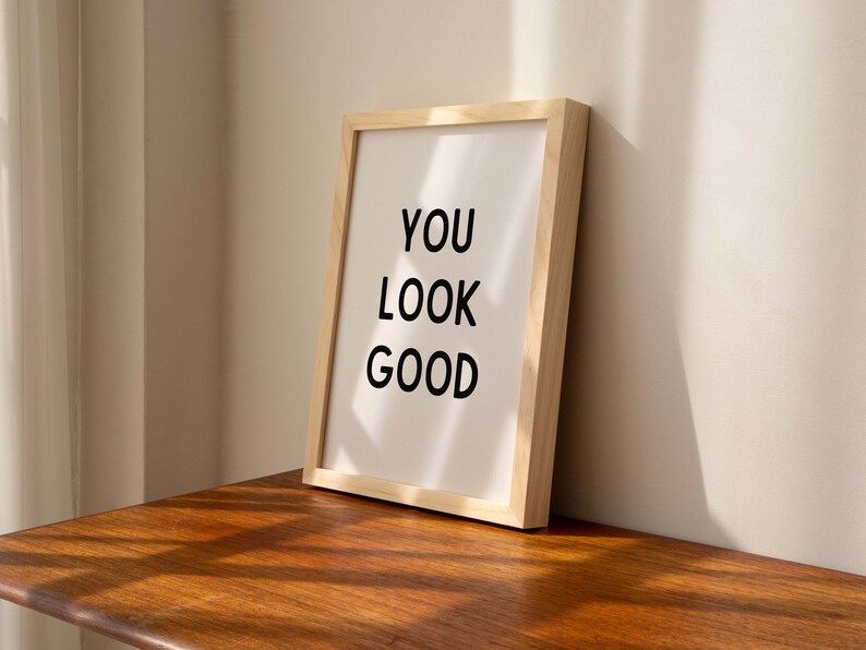 May include: A framed print with the text "YOU LOOK GOOD" in black lettering on a white background. The frame is made of light wood and is sitting on a wooden surface.