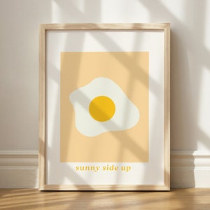 May include: A framed print featuring a yellow egg yolk and white egg white on a peach background. The text "sunny side up" is printed below the egg.