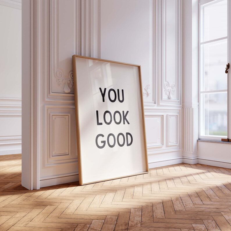 May include: A framed poster with the text "YOU LOOK GOOD" in black lettering on a white background. The poster is leaning against a white wall with ornate molding.
