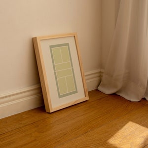 May include: A framed print of a tennis court in green and white colors, leaning against a wall with white trim and wooden floor.