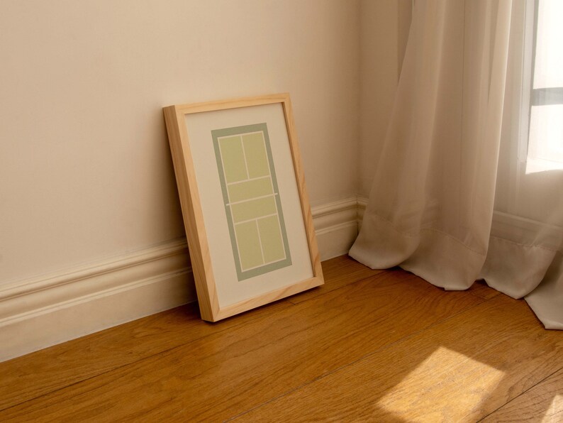 May include: A framed print of a tennis court in green and white colors, leaning against a wall with white trim and wooden floor.