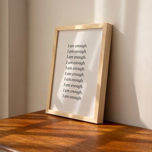 May include: A framed print with the text "I am enough." repeated ten times in black font on a white background. The frame is made of light wood.