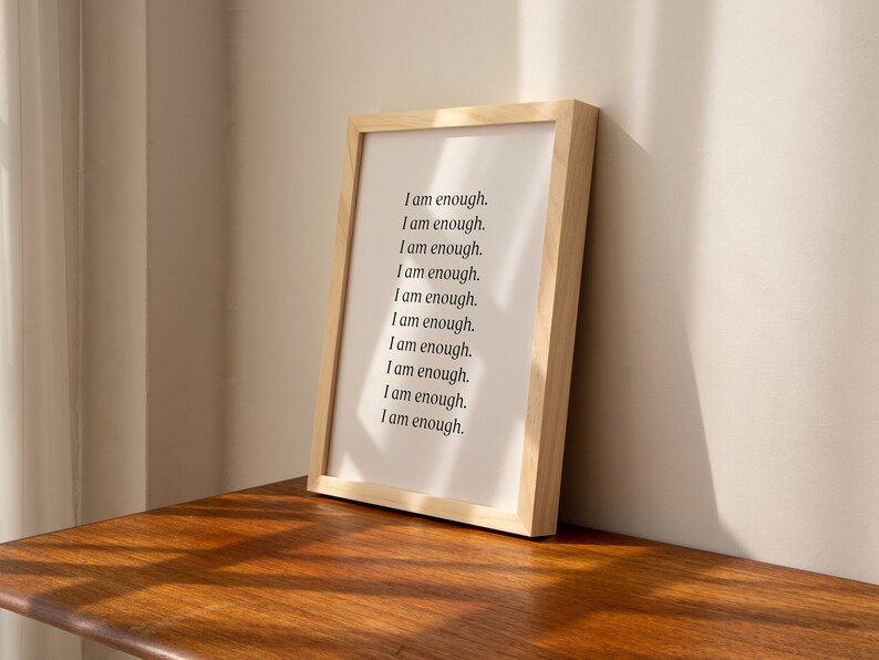 May include: A framed print with the text "I am enough." repeated ten times in black font on a white background. The frame is made of light wood.