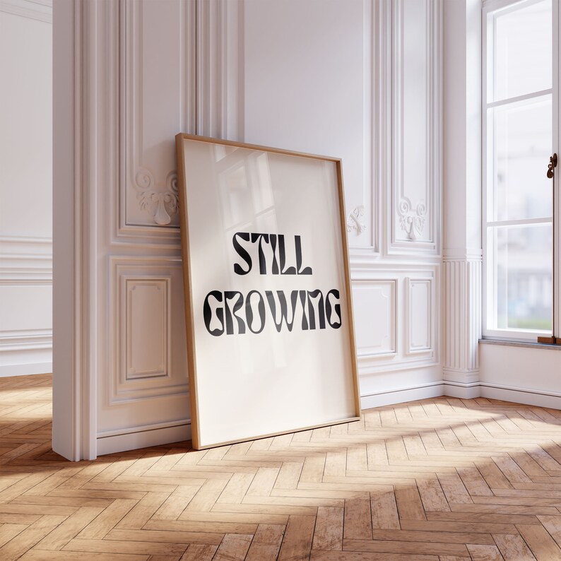 K&ouml;nnte beinhalten: Ein gerahmter Druck mit dem Text "STILL GROWING" in schwarzen Fettdruck auf wei&szlig;em Hintergrund. Der Rahmen ist in hellem Holz gehalten.