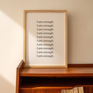 May include: A framed print with the text "I am enough." repeated ten times on a white background. The frame is made of light wood and is sitting on a wooden shelf.