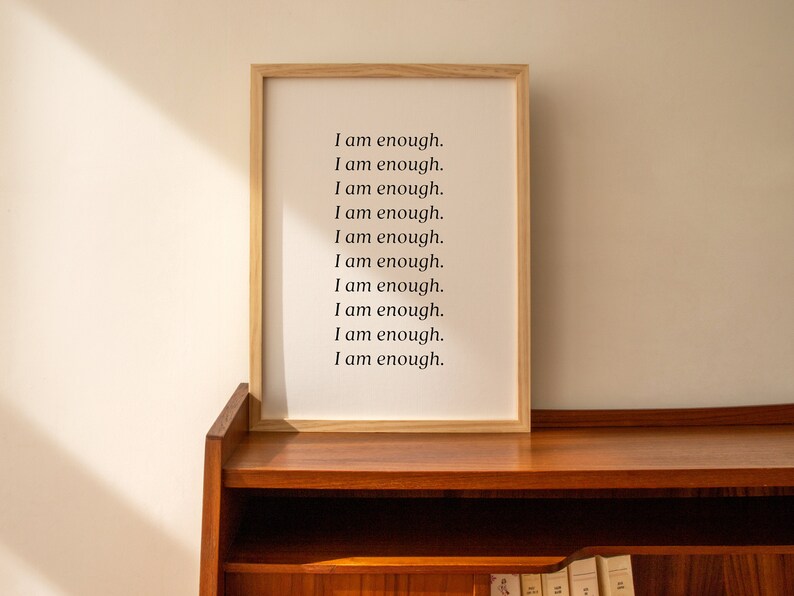 May include: A framed print with the text "I am enough." repeated ten times on a white background. The frame is made of light wood and is sitting on a wooden shelf.
