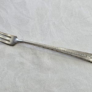 May include: A silver fork with an ornate design on the handle. The fork is on a white fabric background.