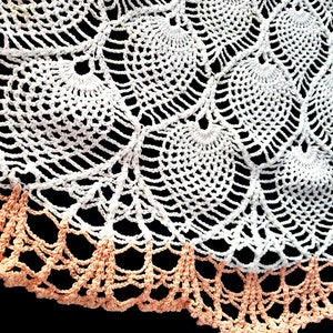 May include: A white crocheted doily with a peach colored crocheted border. The doily has a scalloped edge and a delicate floral pattern.
