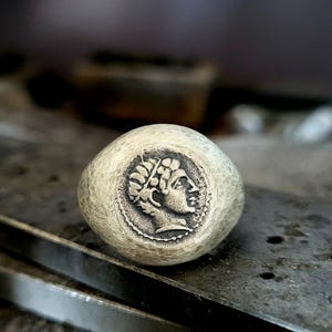 May include: A silver-toned ring with a detailed engraved design of a classical figure's profile within a circular frame. The ring has a textured, hammered appearance. The ring is set on a dark, textured surface, with tools visible in the background.