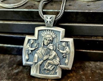 May include: A silver cross pendant featuring a relief of a crowned figure holding a child, flanked by two smaller figures. The pendant is attached to a silver chain with a woven texture, showcasing a dark oxidized finish in the recessed areas.