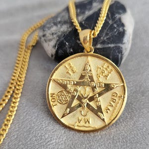 Puede incluir: Un collar con colgante circular dorado que presenta un diseño de pentagrama en relieve. El centro muestra un símbolo de cabeza de cabra, rodeado de caracteres hebreos, la palabra TETRAGRAMMATON y un pequeño hexagrama. El colgante cuelga de una cadena dorada.