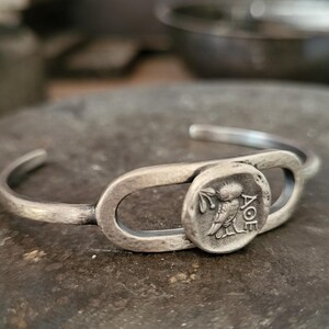 May include: A silver cuff bracelet with an oval centerpiece and a circular coin. The coin features a detailed relief of a bird and Greek lettering. The bracelet has a hammered texture.
