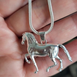 K&ouml;nnte beinhalten: Halskette mit einem silberfarbenen Pferdeanh&auml;nger. Der Anh&auml;nger zeigt ein detailliertes laufendes Pferd. Die Kette hat ein geflochtenes Design. Das Wort "Polished" befindet sich in einem schwarzen Rechteck unten rechts.