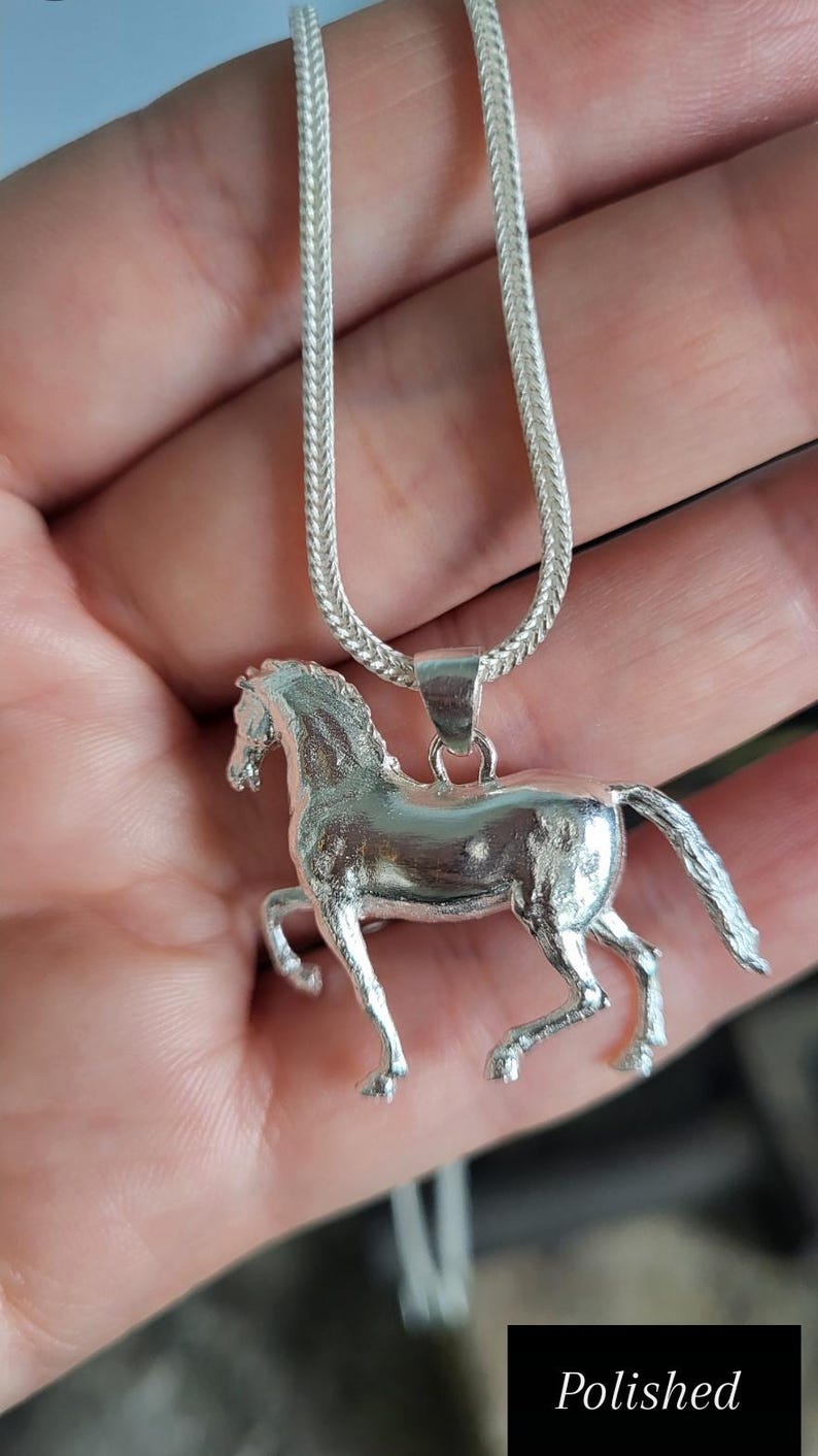 K&ouml;nnte beinhalten: Halskette mit einem silberfarbenen Pferdeanh&auml;nger. Der Anh&auml;nger zeigt ein detailliertes laufendes Pferd. Die Kette hat ein geflochtenes Design. Das Wort "Polished" befindet sich in einem schwarzen Rechteck unten rechts.