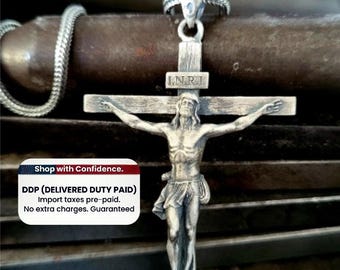 May include: A silver-toned metal crucifix pendant featuring a detailed figure of a person on a cross with the letters I.N.R.I. inscribed on a horizontal plaque above the figure. The pendant is attached to a silver-toned chain with a textured link design.