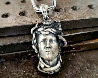 May include: A silver-toned metal pendant featuring a sculpted human face surrounded by multiple intertwined snakes. The pendant is attached to a silver-toned chain and displays a detailed, oxidized finish with dark recessed areas and highlights.