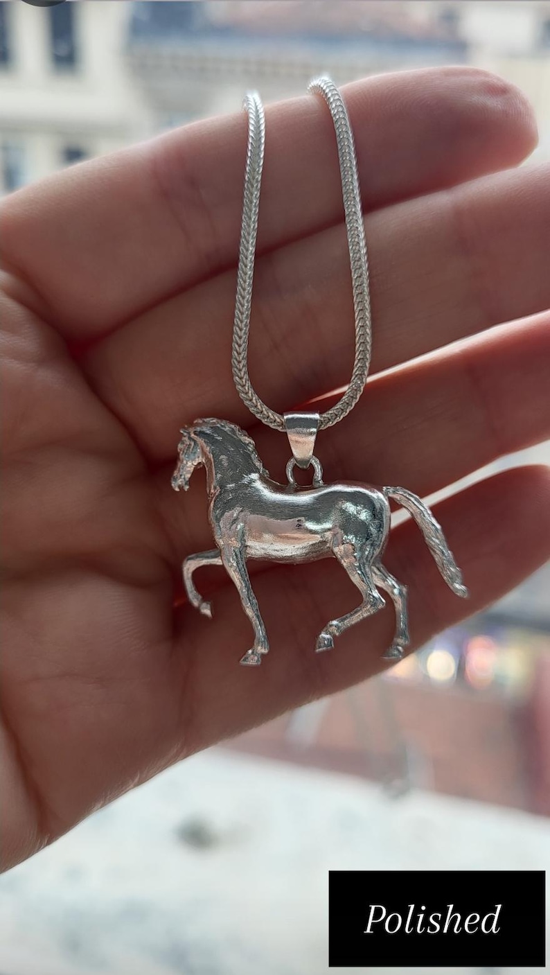 K&ouml;nnte beinhalten: Halskette mit einem silberfarbenen Pferdeanh&auml;nger. Der Anh&auml;nger zeigt ein detailliertes Pferd in einer Laufpose. Die Halskette hat ein geflochtenes Design. Das Wort "Polished" steht in einem schwarzen Feld am unteren Bildrand.