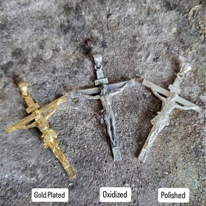 K&ouml;nnte beinhalten: Drei Kruzifix-Anh&auml;nger in vergoldeter, oxidierter und polierter Ausf&uuml;hrung. Jeder Anh&auml;nger zeigt eine detaillierte Darstellung von Jesus am Kreuz. Die Anh&auml;nger werden auf einer strukturierten, grauen Oberfl&auml;che pr&auml;sentiert. Textetiketten kennzeichnen jede Ausf&uuml;hrung.