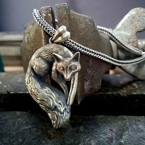 Op de afbeelding: Een zilverkleurige metalen hanger in de vorm van een opgerolde vos, met gedetailleerde vachtstructuur en puntige oren. De hanger is bevestigd aan een gevlochten zilverkleurige ketting en heeft een gepolijste afwerking met donkere accenten.
