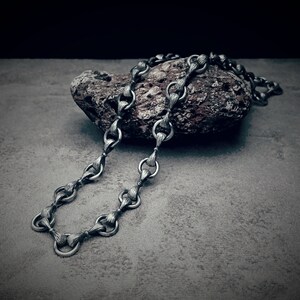 May include: A silver-toned necklace with a textured, ornate design. The necklace is draped over a rough, dark-colored stone, set against a gray background. The chain links have a detailed, almost woven appearance, creating a vintage aesthetic.