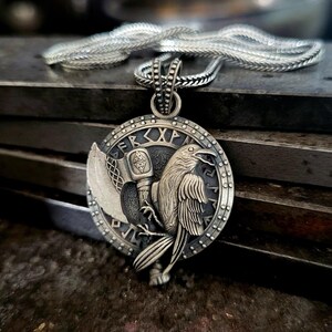 Puede incluir: Collar con colgante de plata con un colgante circular detallado con un diseño de cuervo, martillo y hacha. El colgante tiene runas grabadas y un borde texturizado. La cadena es de estilo trenzado plateado. El collar se muestra sobre una superficie oscura y texturizada.