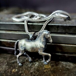 K&ouml;nnte beinhalten: Halskette mit einem silberfarbenen Pferdeanh&auml;nger. Das Pferd ist in einer Laufpose mit detaillierten Merkmalen dargestellt. Die Kette ist ebenfalls silberfarben. Das Schmuckst&uuml;ck liegt auf einer strukturierten Oberfl&auml;che.