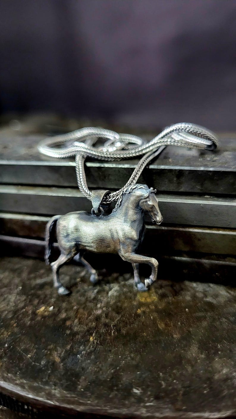 K&ouml;nnte beinhalten: Halskette mit einem silberfarbenen Pferdeanh&auml;nger. Das Pferd ist in einer Laufpose mit detaillierten Merkmalen dargestellt. Die Kette ist ebenfalls silberfarben. Das Schmuckst&uuml;ck liegt auf einer strukturierten Oberfl&auml;che.