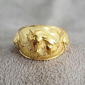May include: A gold-coloured ring featuring a design of three horse heads. The ring is on a grey fuzzy surface.