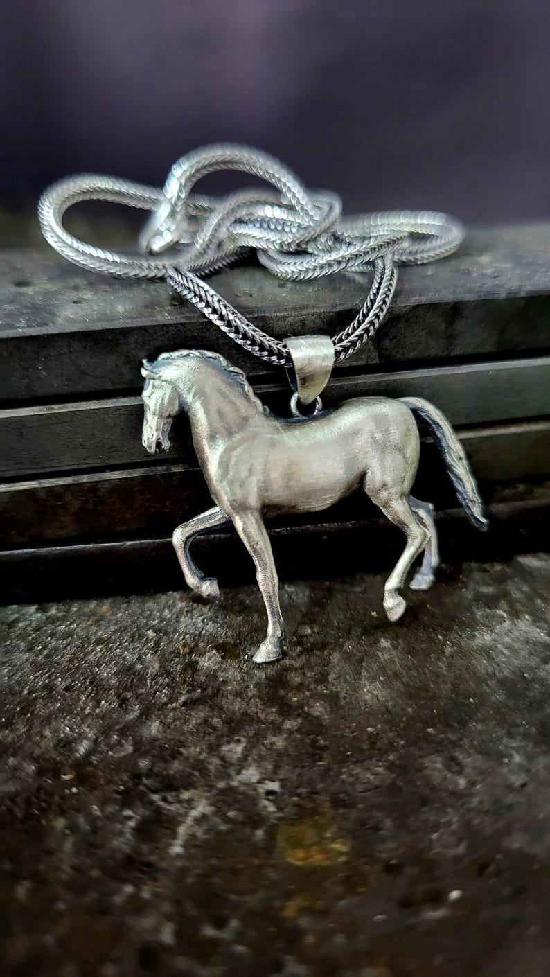 K&ouml;nnte beinhalten: Halskette mit einem silbernen Pferdeanh&auml;nger. Der Anh&auml;nger ist eine detaillierte Skulptur eines Pferdes in einer Laufpose. Die Halskette hat ein geflochtenes Design. Der Pferdeanh&auml;nger und die Kette sind aus gl&auml;nzendem Metall.
