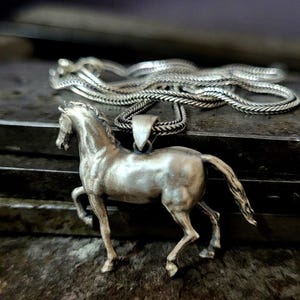 K&ouml;nnte beinhalten: Halskette mit silberfarbenem Pferdeanh&auml;nger. Der Anh&auml;nger ist eine detaillierte Skulptur eines galoppierenden Pferdes. Die Halskette hat ein geflochtenes Design. Der Schmuck wird auf einer strukturierten, dunklen Oberfl&auml;che pr&auml;sentiert.