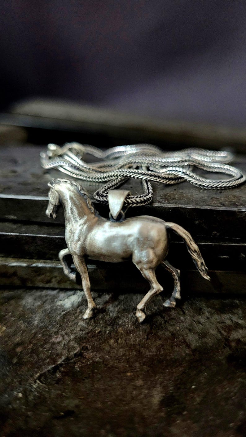 K&ouml;nnte beinhalten: Halskette mit silberfarbenem Pferdeanh&auml;nger. Der Anh&auml;nger ist eine detaillierte Skulptur eines galoppierenden Pferdes. Die Halskette hat ein geflochtenes Design. Der Schmuck wird auf einer strukturierten, dunklen Oberfl&auml;che pr&auml;sentiert.