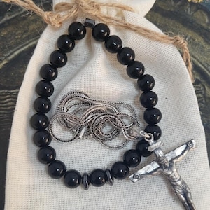 K&ouml;nnte beinhalten: Ein schwarzes Perlenarmband und eine silberne Halskette mit Kreuzanh&auml;nger liegen auf einem kleinen wei&szlig;en Stoffbeutel. Der Beutel ist mit Naturfaden gebunden. Der Hintergrund hat goldene dekorative Elemente.