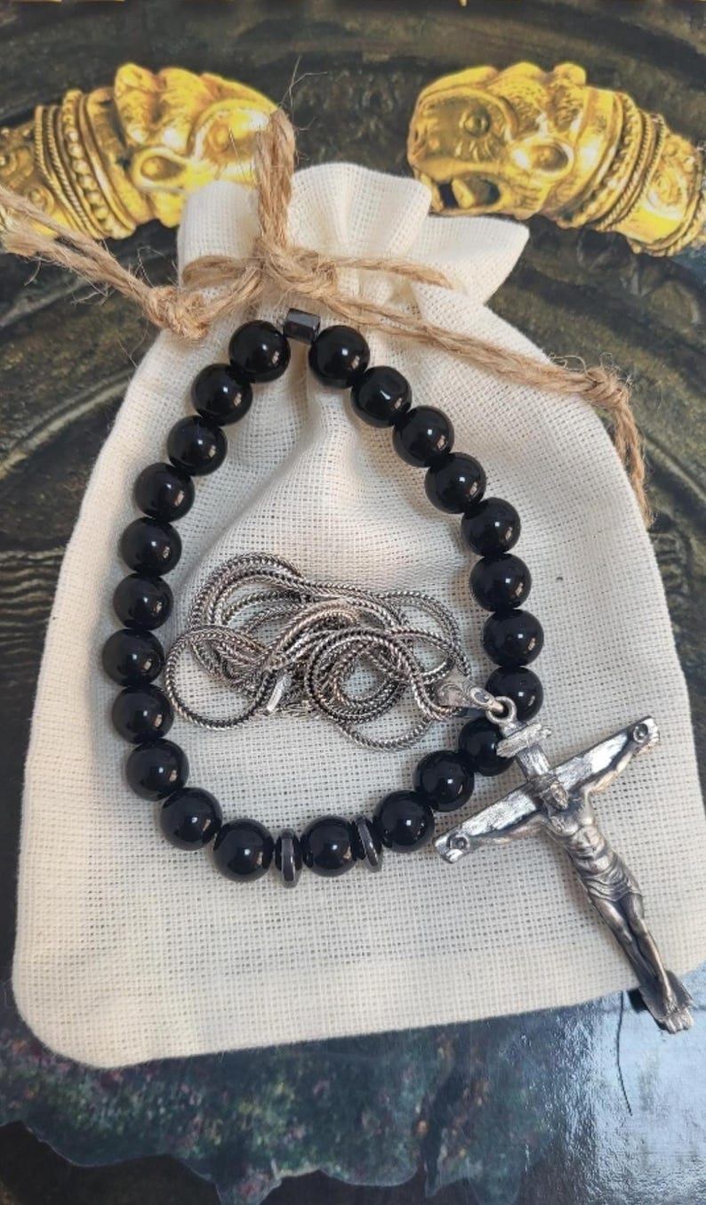 K&ouml;nnte beinhalten: Ein schwarzes Perlenarmband und eine silberne Halskette mit Kreuzanh&auml;nger liegen auf einem kleinen wei&szlig;en Stoffbeutel. Der Beutel ist mit Naturfaden gebunden. Der Hintergrund hat goldene dekorative Elemente.