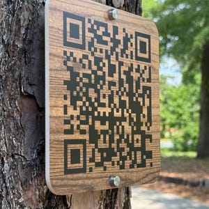 May include: A rectangular wooden sign with a QR code is attached to a tree. The sign has a wood grain pattern and is secured with two silver screws. The background shows a blurred view of a park.