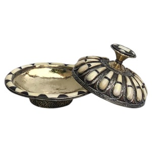 This is a decorative small dish made from a combination of copper and bone. Brass gives it a warm, reflective surface with a hammered texture, while bone inlays add a decorative touch similar to that of ivory, providing a contrast to the metal.
