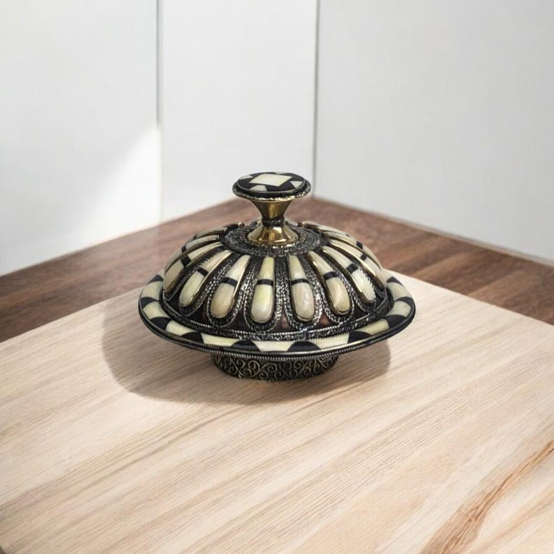 This is a decorative small dish made from a combination of copper and bone. Brass gives it a warm, reflective surface with a hammered texture, while bone inlays add a decorative touch similar to that of ivory, providing a contrast to the metal.