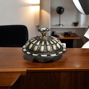 This is a decorative small dish made from a combination of copper and bone. Brass gives it a warm, reflective surface with a hammered texture, while bone inlays add a decorative touch similar to that of ivory, providing a contrast to the metal.