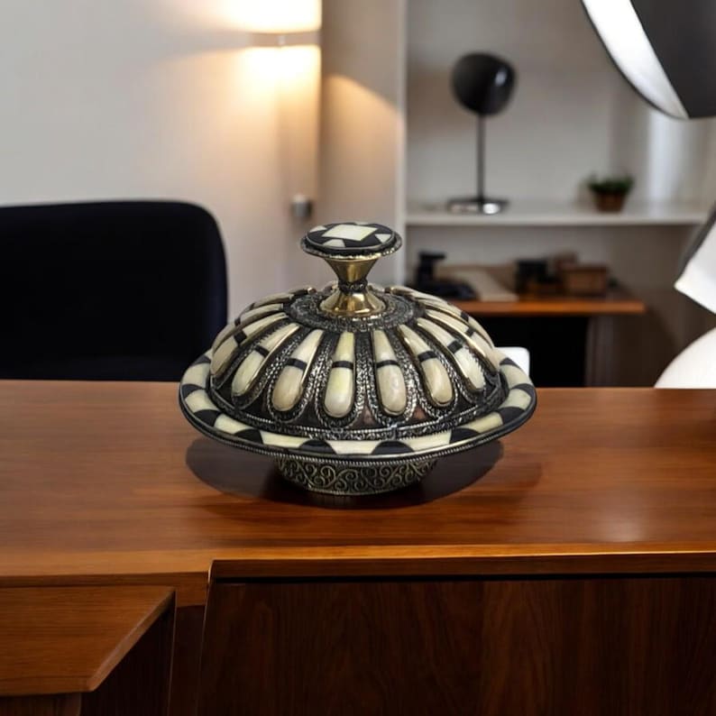 This is a decorative small dish made from a combination of copper and bone. Brass gives it a warm, reflective surface with a hammered texture, while bone inlays add a decorative touch similar to that of ivory, providing a contrast to the metal.
