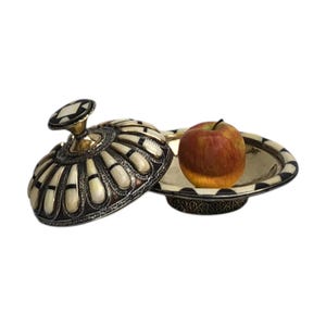 This is a decorative small dish made from a combination of copper and bone. Brass gives it a warm, reflective surface with a hammered texture, while bone inlays add a decorative touch similar to that of ivory, providing a contrast to the metal.