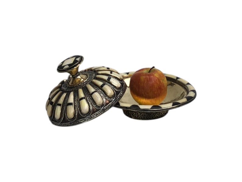 This is a decorative small dish made from a combination of copper and bone. Brass gives it a warm, reflective surface with a hammered texture, while bone inlays add a decorative touch similar to that of ivory, providing a contrast to the metal.