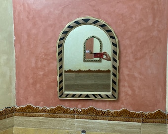 May include: Arched mirror with a black and gold geometric border, reflecting a room with a smaller mirror. The mirror is mounted on a textured, reddish-pink wall. The bottom of the wall has a decorative border.