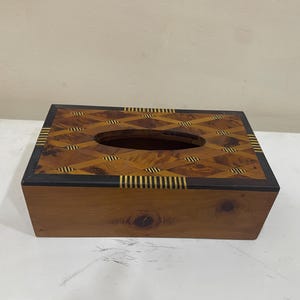 Puede incluir: Caja de pañuelos rectangular de madera con un diseño geométrico en la tapa. La tapa presenta un patrón de diamantes en tonos marrones y dorados, con un borde marrón oscuro y una abertura ovalada.