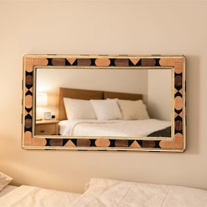 Peut inclure: Miroir rectangulaire avec un cadre en bois décoratif. Le cadre présente un motif géométrique avec des accents de bois noirs, bruns et de couleur claire. Le miroir est accroché à un mur beige, reflétant une scène de chambre.
