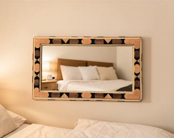 May include: Rectangular mirror with a decorative wooden frame. The frame features a geometric pattern with black, brown, and light-colored wood accents. The mirror is hanging on a beige wall, reflecting a bedroom scene.