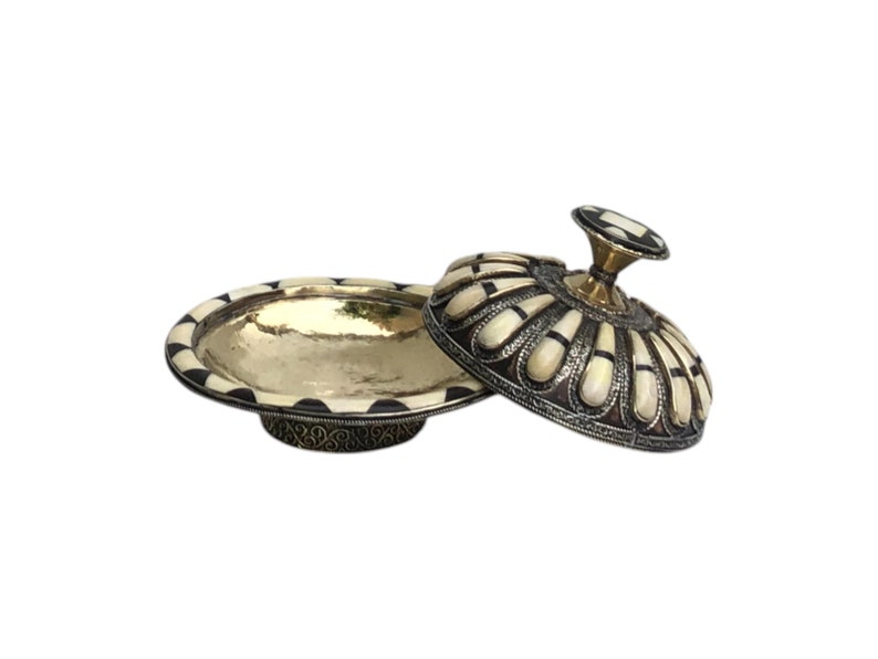 This is a decorative small dish made from a combination of copper and bone. Brass gives it a warm, reflective surface with a hammered texture, while bone inlays add a decorative touch similar to that of ivory, providing a contrast to the metal.