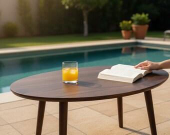 Puede incluir: Una mesa de centro ovalada de madera oscura con cuatro patas cónicas. Sobre la mesa hay un vaso transparente con zumo de naranja y cubitos de hielo, junto a un libro blanco abierto con páginas visibles. La mesa está situada en un patio pavimentado.