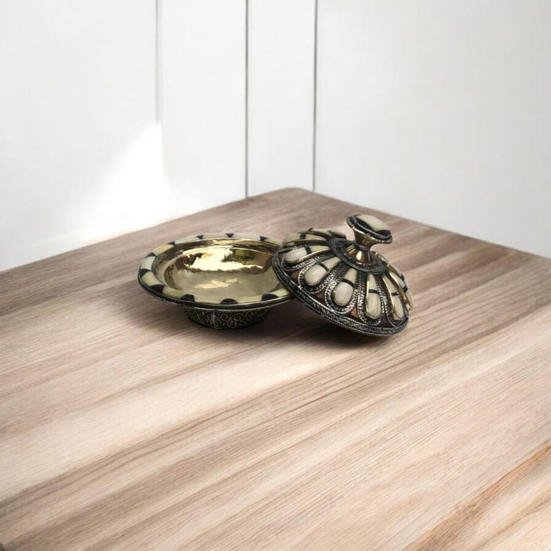 This is a decorative small dish made from a combination of copper and bone. Brass gives it a warm, reflective surface with a hammered texture, while bone inlays add a decorative touch similar to that of ivory, providing a contrast to the metal.